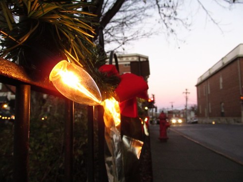 A close-up of festive lights on a fence, with a sunset in the background and a street scene visible.