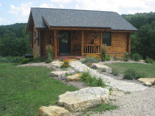 A cozy log cabin surrounded by colorful flowers and greenery, with a stone pathway leading to the entrance.
