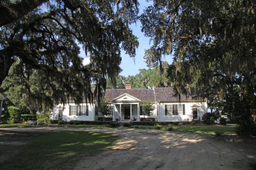 A white house with a porch, surrounded by trees and greenery, under a clear blue sky.