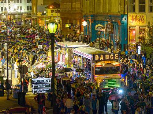 A vibrant street scene at night with a colorful bus, crowds, and umbrellas during a festive event in Galveston.