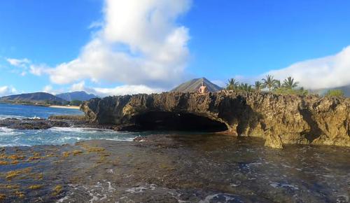 A rocky coastline with a natural arch, clear blue sky, and a person standing on the rock formation near the water.