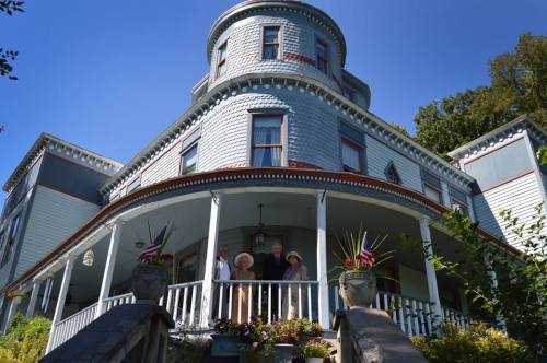 A historic blue house with a round turret, featuring a porch and four people standing on the steps, surrounded by greenery.