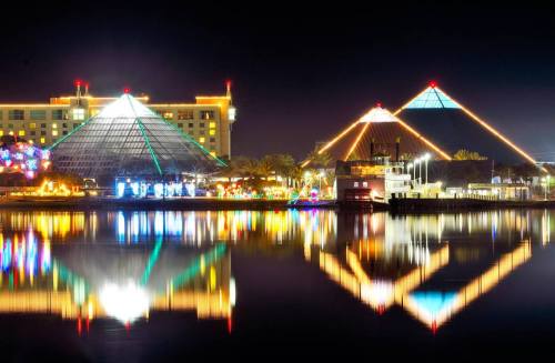 Night view of illuminated pyramid-shaped buildings reflecting in water, with colorful lights and a serene atmosphere.