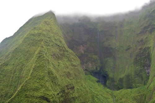 Lush green mountain peaks shrouded in mist, with a waterfall cascading down the rocky cliff face.