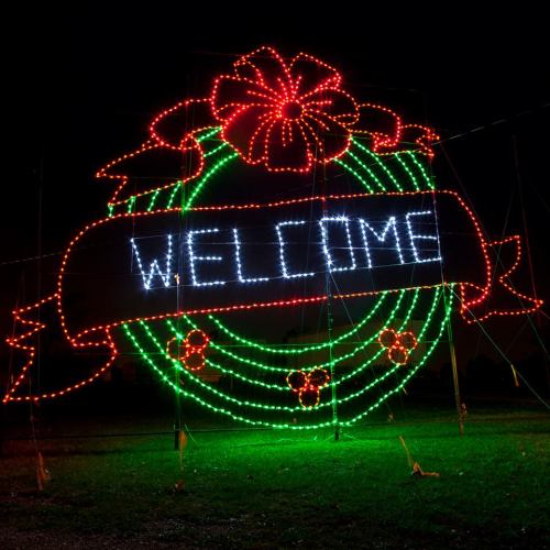 A brightly lit holiday wreath with a red bow and the word "WELCOME" in white lights against a dark background.