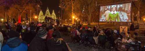 A crowd watches a movie outdoors on a large screen, surrounded by festive lights and trees at night.