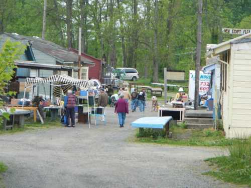 A bustling outdoor market scene with people browsing stalls and shops amidst greenery and rustic buildings.