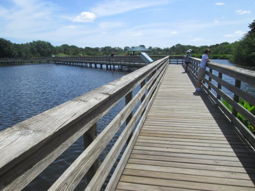 A wooden boardwalk extends over a calm lake, with people enjoying the view under a clear blue sky.