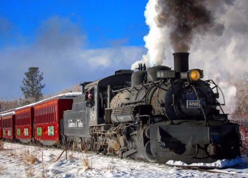 A vintage steam train billows smoke while traveling through a snowy landscape, with red passenger cars behind it.