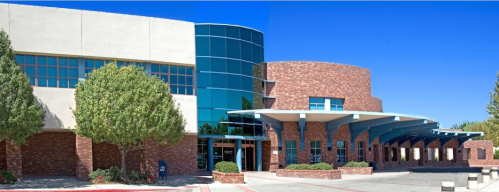 Modern brick and glass building with a curved entrance and landscaped trees under a clear blue sky.