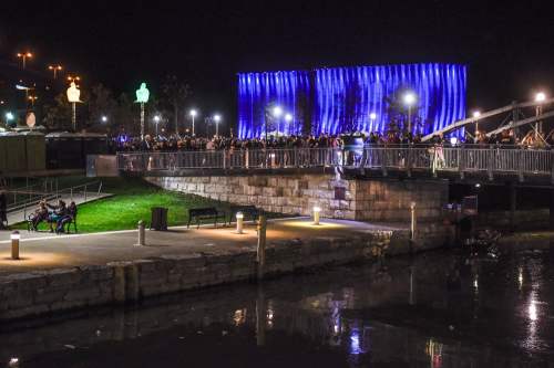 A vibrant night scene by a river, featuring a crowd near a blue-lit building and illuminated pathways.