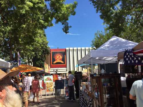 A bustling outdoor market with colorful tents, people browsing, and trees under a clear blue sky.