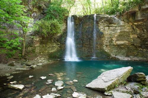 A serene waterfall cascades into a clear pool, surrounded by lush greenery and rocky terrain.