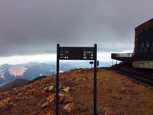 Signpost on a mountain with cloudy skies, indicating directions and distances, near a building and rocky terrain.