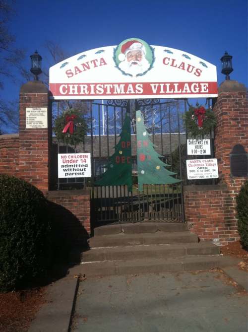 Entrance gate to Santa Claus Christmas Village, decorated with wreaths and a Christmas tree, with signs about visiting hours.