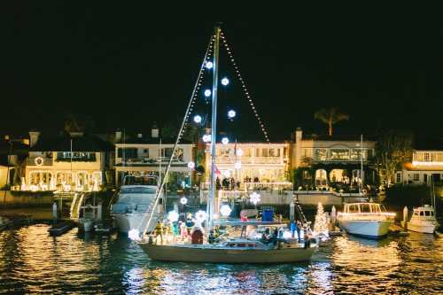 A decorated sailboat with lights floats on a calm waterway, surrounded by festive homes at night.