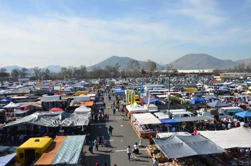 A bustling outdoor market filled with colorful tents and crowds, set against a backdrop of mountains and clear skies.