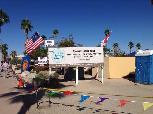 Sign for a monthly market event with palm trees, flags, and colorful decorations in a sunny outdoor setting.