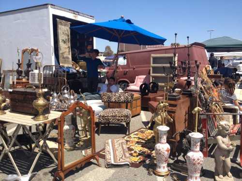 A bustling flea market scene with various antiques, furniture, and a vendor under a blue umbrella.