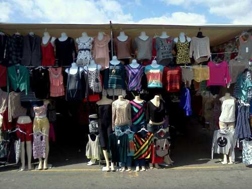 A clothing stall displaying various tops and skirts on mannequins under a canopy, with a blue sky in the background.