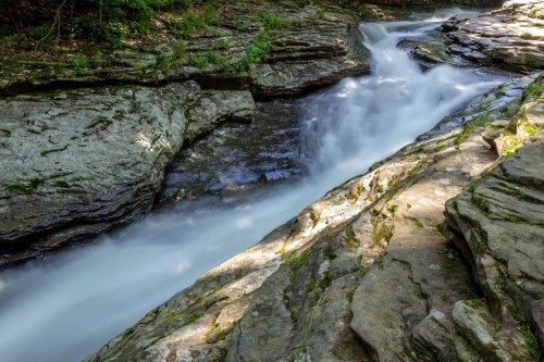 A serene river flows over smooth rocks, surrounded by lush greenery and dappled sunlight.