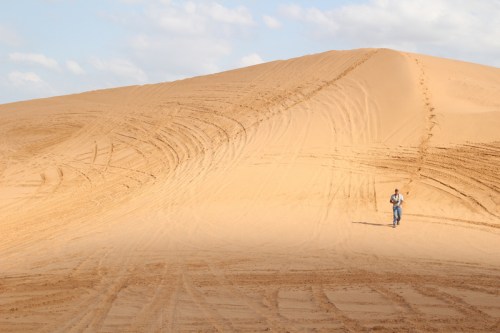 A lone person walks across a vast sandy dune under a partly cloudy sky, with tire tracks visible in the sand.