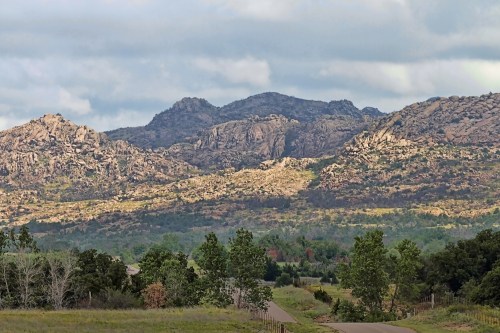 A scenic view of rocky mountains under a cloudy sky, with a winding road and green trees in the foreground.