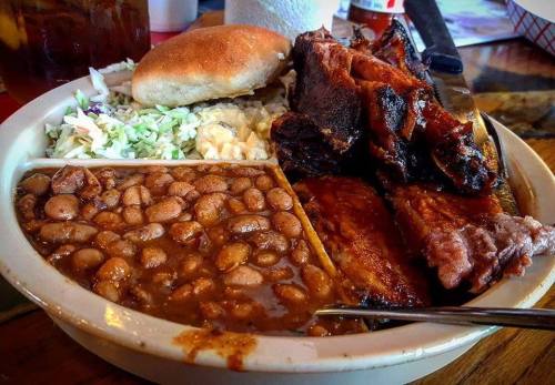 A plate of barbecue featuring ribs, brisket, baked beans, coleslaw, and a bun, served with a fork.