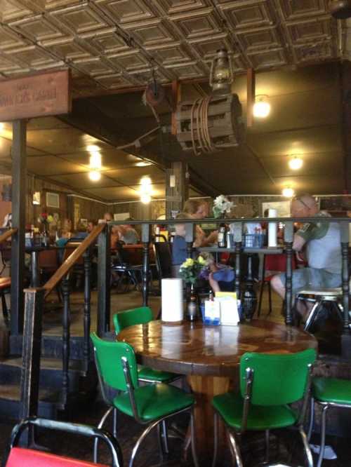 A cozy restaurant interior with wooden tables, green chairs, and patrons enjoying their meals under warm lighting.