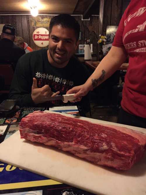 A man gives a thumbs-up next to a large piece of raw meat on a cutting board in a rustic setting.