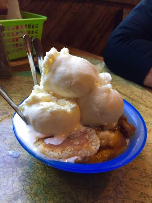 A blue bowl filled with warm peach cobbler topped with three scoops of vanilla ice cream and two silver spoons.