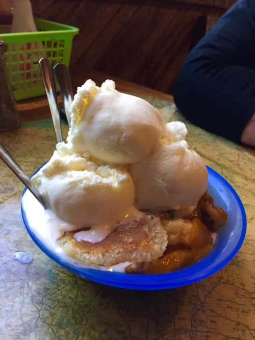 A blue bowl filled with warm peach cobbler topped with three scoops of vanilla ice cream and two silver spoons.