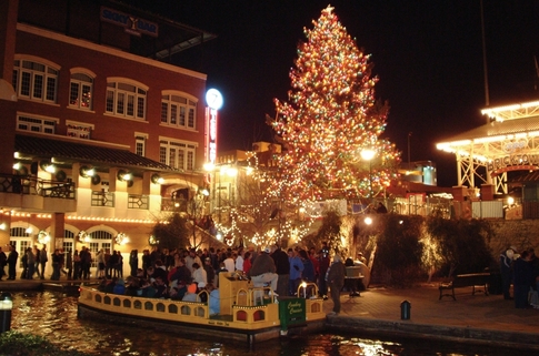 A festive scene at night with a brightly lit Christmas tree and a boat on a canal, surrounded by holiday decorations.