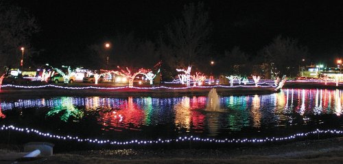 Colorful holiday lights reflect on a pond at night, with illuminated trees and a fountain creating a festive atmosphere.