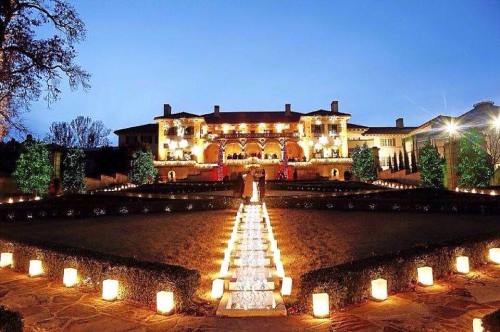 A grand mansion illuminated at dusk, with a pathway lined by glowing lanterns and lush greenery.