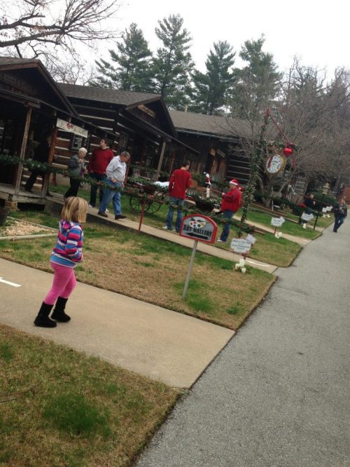 A child in pink pants walks past festive cabins with people decorating for the holidays in a wooded area.