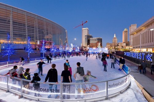 A bustling ice skating rink surrounded by festive lights and a city skyline at dusk.