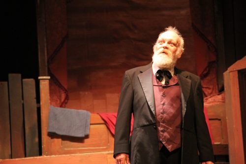 An elderly man in formal attire stands on stage, looking upward with a thoughtful expression, against a rustic backdrop.