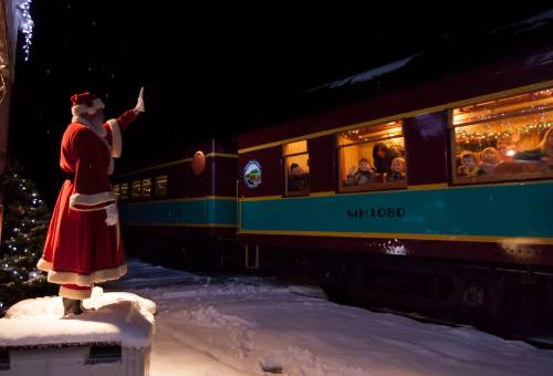 A Santa figure waves to passengers on a festive train, surrounded by snow and holiday decorations.
