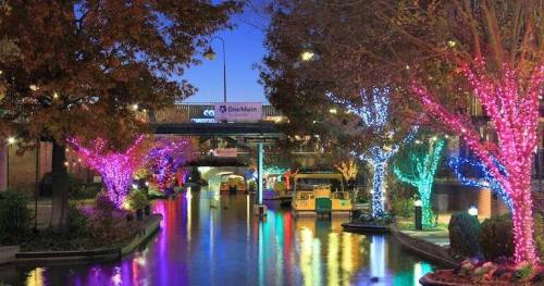 Colorful lights adorn trees along a serene canal at dusk, creating a festive and inviting atmosphere.