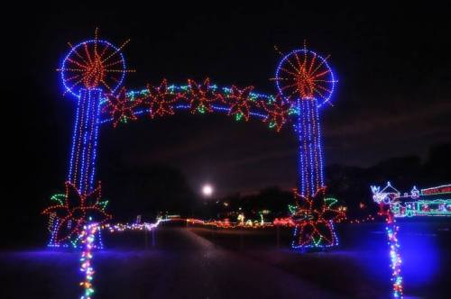 Colorful holiday lights form an archway, illuminating a path with festive decorations against a night sky.