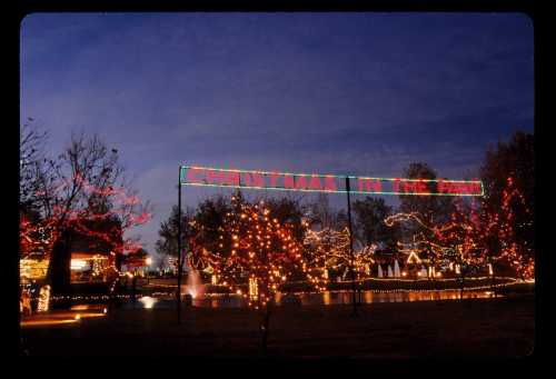 Colorful holiday lights illuminate a park at dusk, featuring a sign that reads "Christmas in the Park."