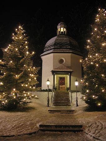 A cozy, snow-covered chapel surrounded by illuminated Christmas trees at night.