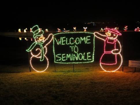 Two illuminated snowmen holding a sign that reads "Welcome to Seminole" at night, with festive lights in the background.