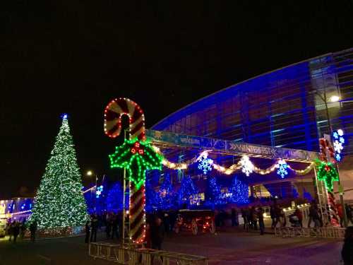 A festive scene featuring a large Christmas tree, colorful lights, and holiday decorations outside a building at night.