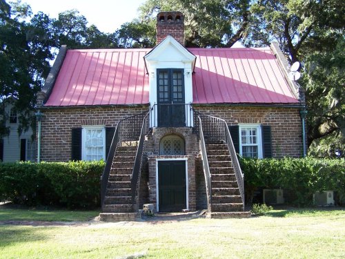 A charming brick house with a red metal roof, featuring a central door and two staircases leading to the entrance.