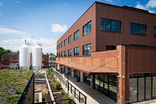 Modern building with large windows, a green roof, and silos nearby, set against a blue sky with scattered clouds.