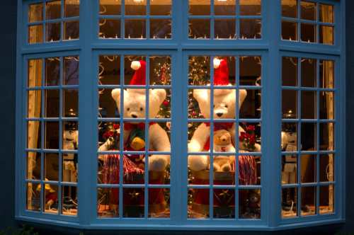 Two large teddy bears in Santa hats sit in a festive window, surrounded by Christmas decorations and a tree.