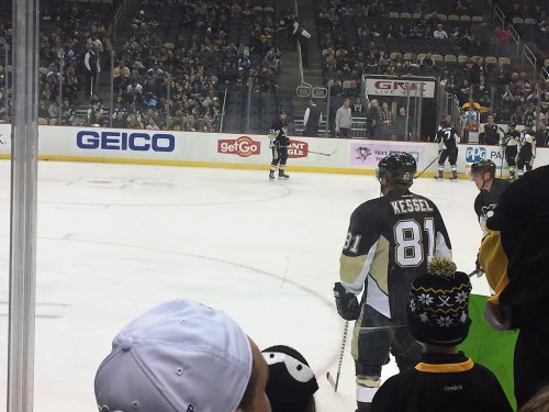 A hockey player in a black and gold jersey stands on the ice, with fans and players visible in the background.