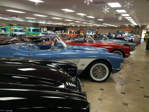 A row of classic cars in a showroom, featuring blue, red, and black convertibles under bright lights.